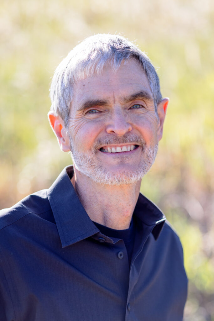 An older man with short gray hair and a beard smiles outdoors, wearing a dark collared shirt. The sunlight and greenery blur behind him as if he's just left a Colorado Choir rehearsal.