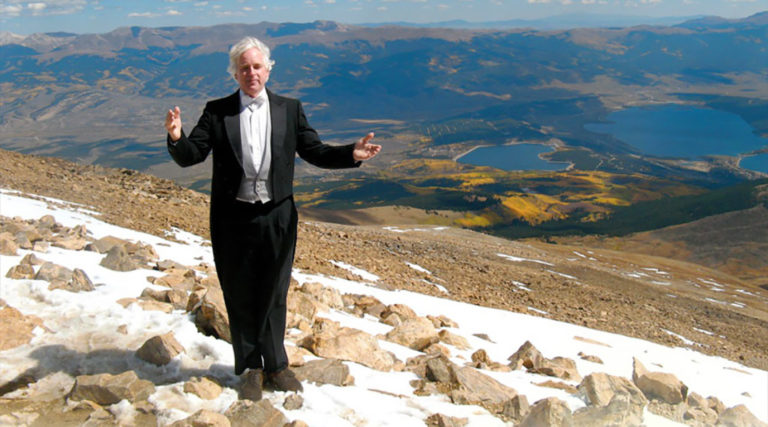 A man in a tuxedo, perhaps a member of the Colorado Choir, stands on a rocky, snow-dusted mountain slope with lakes and mountains visible in the distance under a clear sky.