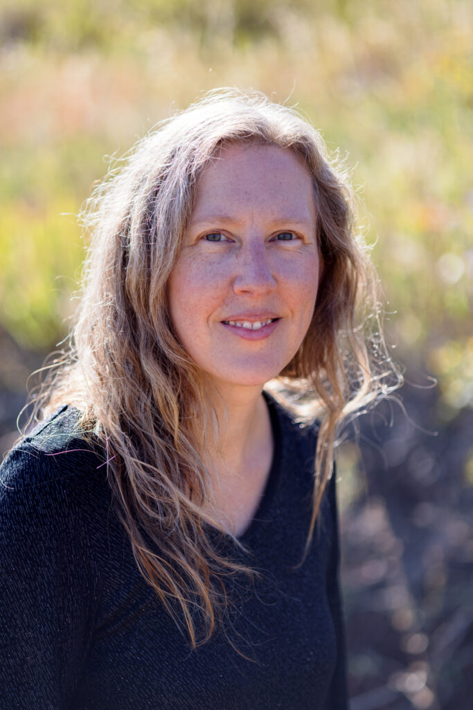 A woman with long, wavy, light brown hair and a black top stands outdoors in natural sunlight with a blurred grassy background, evoking the serene presence of a Colorado Choir member.