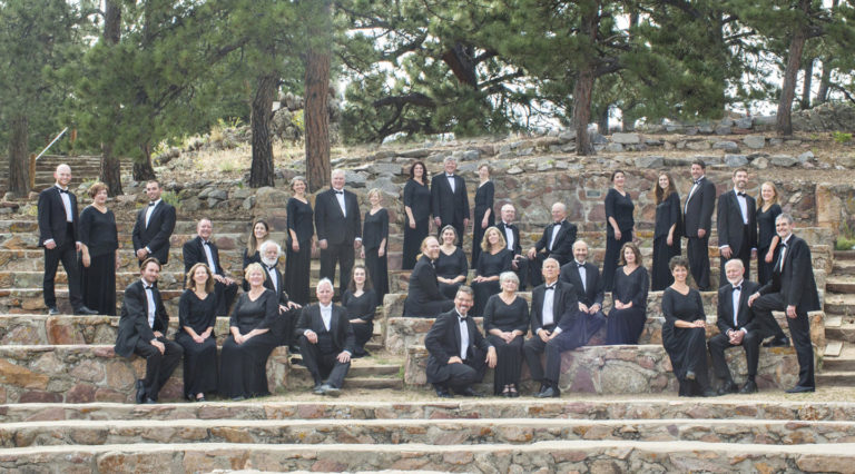 Members of the Colorado Choir, dressed in formal black attire, pose together on stone steps in an outdoor amphitheater surrounded by trees.