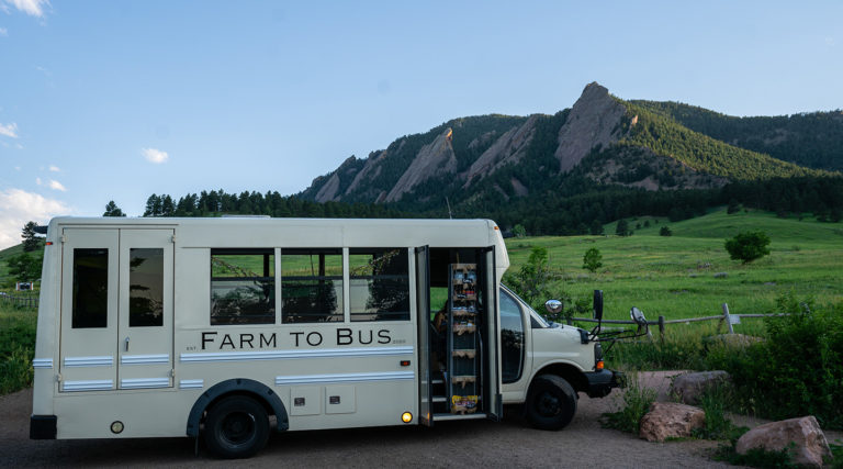 A cream-colored bus labeled "FARM TO BUS" is parked on a dirt road, with shelves visible inside. Rocky mountains and green fields stretch out beneath a clear sky, as if awaiting the arrival of the Colorado Choir for an open-air performance.