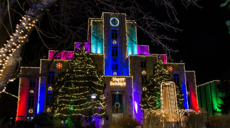 A building is decorated with colorful lights and holiday wreaths at night, with “Happy Holidays” displayed above the entrance, as the Colorado Choir performs and nearby trees are wrapped in sparkling string lights.