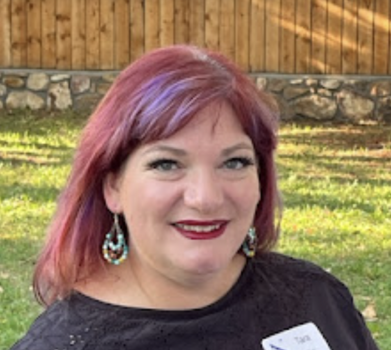 A woman with red hair and beaded earrings smiles outdoors, wearing a black top, a name tag, and representing the Colorado Choir. A stone wall and wooden fence are visible in the background.