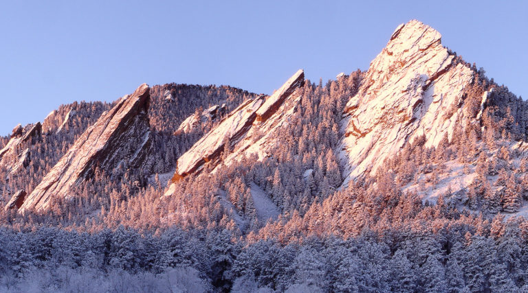 Snow-covered flatiron rock formations rise above a forest of pine trees, illuminated by soft pinkish sunlight against a clear blue sky—nature’s harmony echoing like a Colorado Choir.