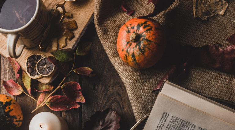 A cup of coffee, a candle, a pumpkin, dried leaves, and an open book arranged on a dark wooden table with burlap fabric set the perfect autumn mood—ideal for cozy moments or listening to the Colorado Choir.
