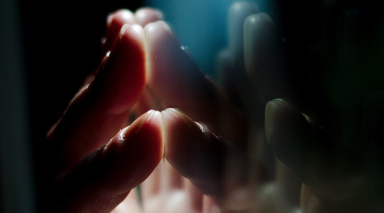 A close-up of fingers touching a reflective glass surface, with the hand's reflection visible and soft lighting highlighting the fingertips, reminiscent of the gentle harmony of a Colorado Choir.