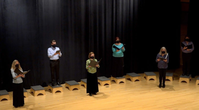 Six members of the Colorado Choir stand on a stage in front of a black curtain, spaced apart, holding folders or books and wearing masks.