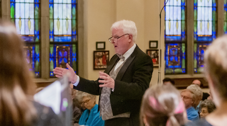 An older man in formal attire stands with arms outstretched, appearing to conduct the Colorado Choir before an audience in a church with stained glass windows.
