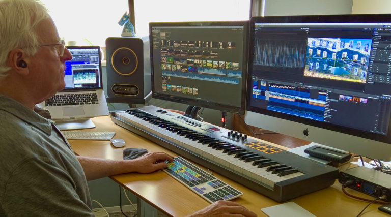 A man works at a desk with two monitors, a keyboard, and a laptop, editing video and audio files from the Colorado Choir in a home studio setup.