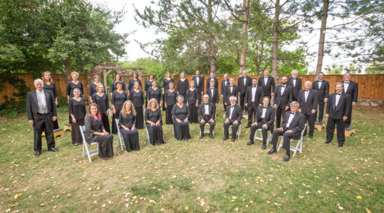 A large Colorado Choir group dressed in formal black attire poses outdoors in front of trees and a wooden fence, with some seated and others standing.