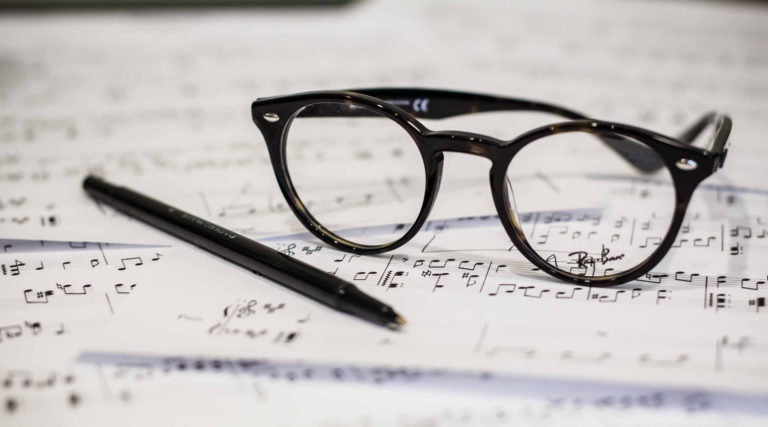 A pair of round eyeglasses and a black pen rest on scattered sheets of handwritten music notation, awaiting the next rehearsal with the Colorado Choir.