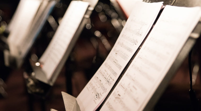 Close-up of several music stands holding sheets of printed musical notation in a dimly lit setting, likely in preparation for a Colorado Choir performance or rehearsal.