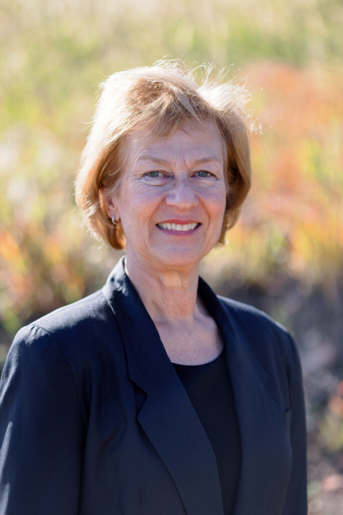 An older woman with short blonde hair, wearing a black blazer, stands outdoors in front of a blurred background of grass and plants, reminiscent of a Colorado Choir concert setting.