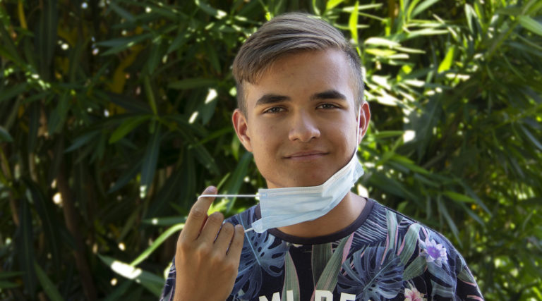 A young man standing outdoors lowers his surgical face mask below his chin while looking at the camera, with green foliage in the background—perhaps pausing between Colorado Choir rehearsals.