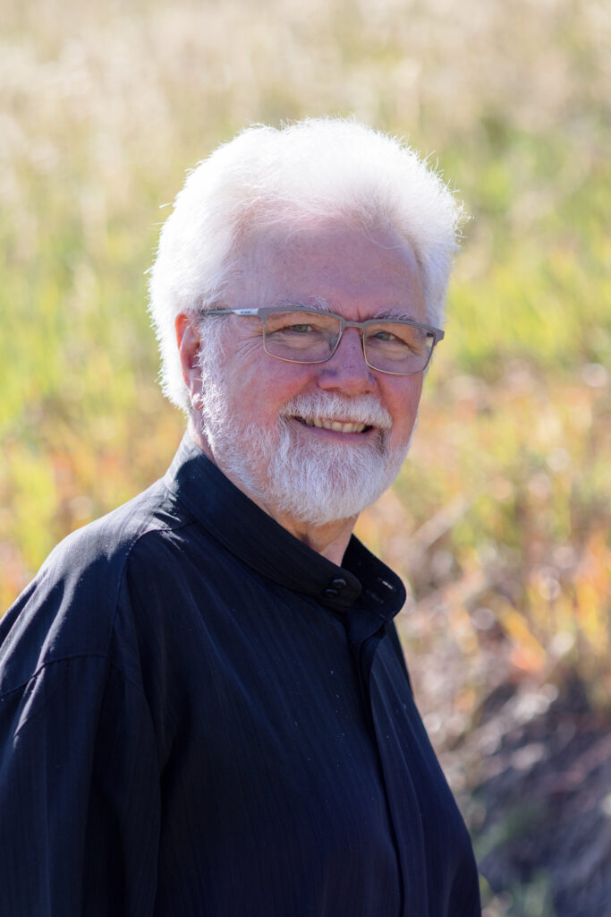 An older man with white hair, a beard, and glasses, wearing a black shirt, stands outdoors in front of a blurred grassy background, smiling at the camera—possibly a proud member of the Colorado Choir.