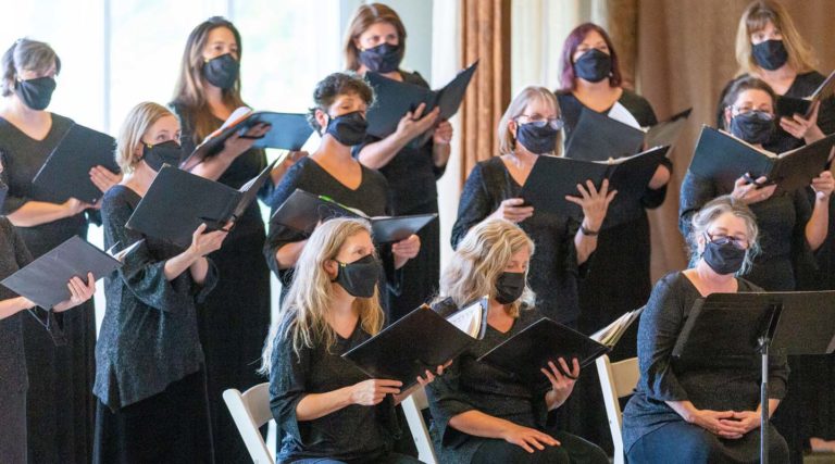 Members of the Colorado Choir, dressed in black masks and attire, sing from sheet music—some standing, others seated—creating a powerful choral performance.