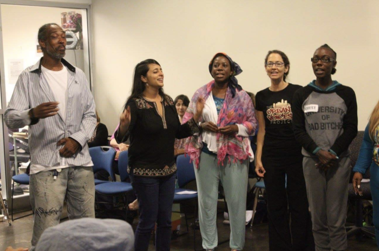 Five people stand in a row at the front of a room, appearing to sing or speak, as if performing with a Colorado Choir, with seated individuals and a glass-walled office space in the background.