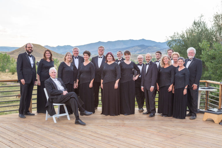 A Colorado Choir group of sixteen people in black formal attire poses on a wooden deck outdoors with mountains and trees in the background.