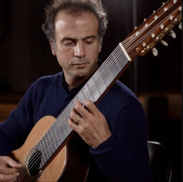 A man in a blue sweater plays a multi-string classical guitar, focusing intently on finger placement, as the Colorado Choir performs in the background.