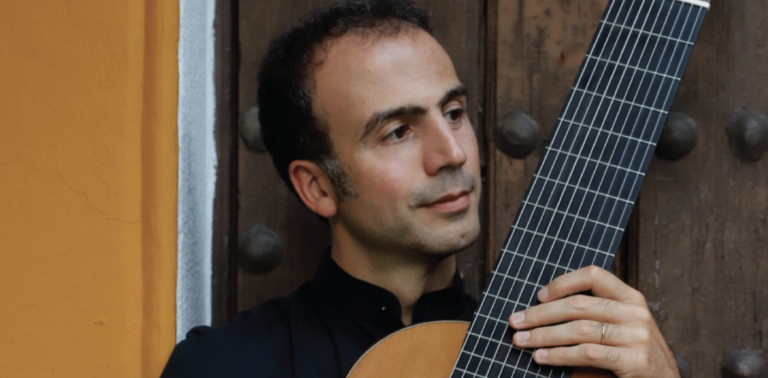 A man holding a classical guitar stands in front of a wooden door with metal studs and an orange wall, ready to perform with the Colorado Choir.