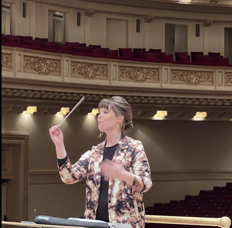 A woman in a patterned blazer conducts in an empty concert hall, standing at a podium with a baton raised in her right hand, preparing for the Colorado Choir’s next performance.