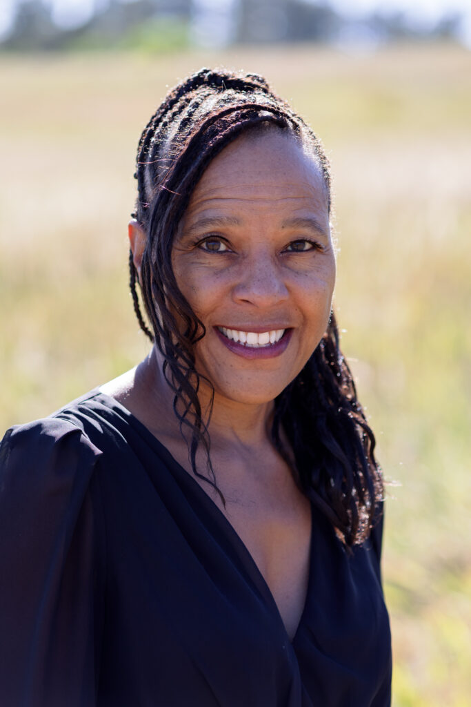 A woman with braided hair, wearing a black top, stands outdoors in a sunlit Colorado Choir field, smiling at the camera.