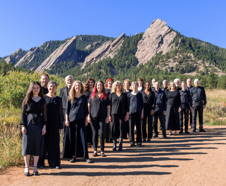 A group of people dressed in black stand in a line on a dirt path with rocky mountains and trees in the background under a clear blue sky.