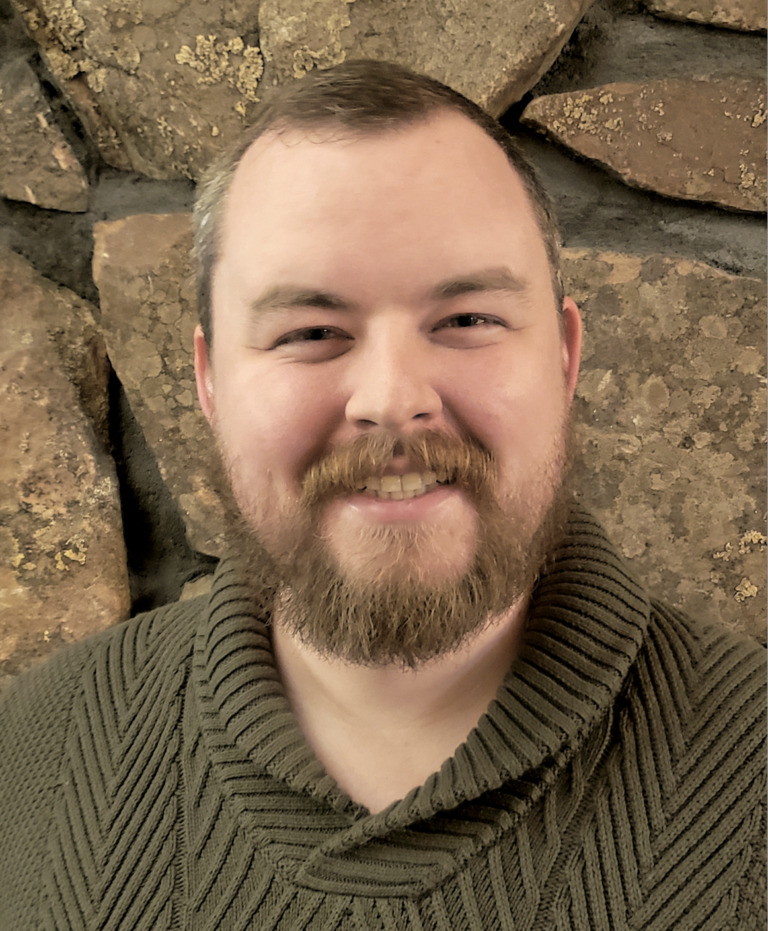 A bearded man wearing a green sweater smiles in front of a stone wall, proudly representing the Colorado Choir.