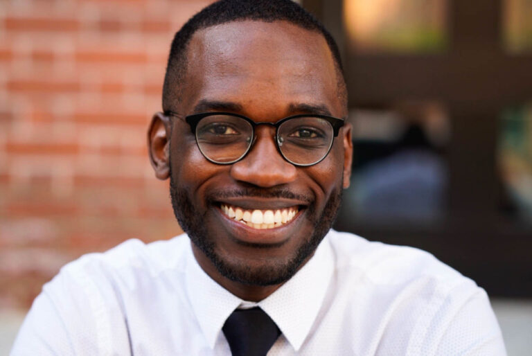 A man wearing glasses, a white shirt, and a black tie smiles at the camera. The blurred background and brick wall suggest he might be posing after a Colorado Choir performance.
