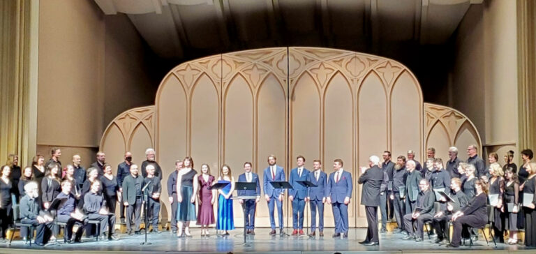 Members of the Colorado Choir and soloists stand on stage in formal attire, facing the audience, with a conductor and music stands arranged before a decorative backdrop.