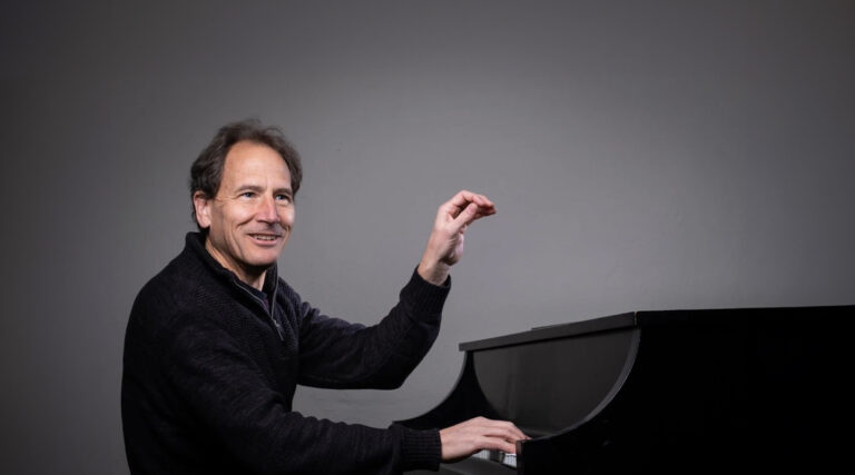 A man sits at a black grand piano, smiling, with one hand on the keys and the other raised in a conducting gesture, as if leading the Colorado Choir, against a plain gray background.