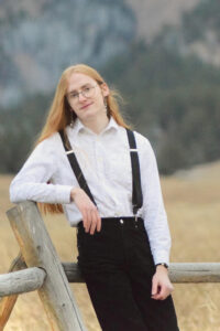 Zahnay Marks, with long red hair, wearing glasses, a white shirt, black suspenders, and black pants, leans on a wooden fence against a natural landscape backdrop.