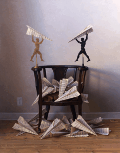 A wooden chair sits on a wooden floor, surrounded by paper airplanes made from sheet music; two small winged figurines stand atop the chair holding airplanes.