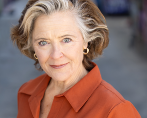 Older woman with short gray and brown hair, wearing gold hoop earrings and an orange collared shirt, looking directly at the camera with a slight smile.