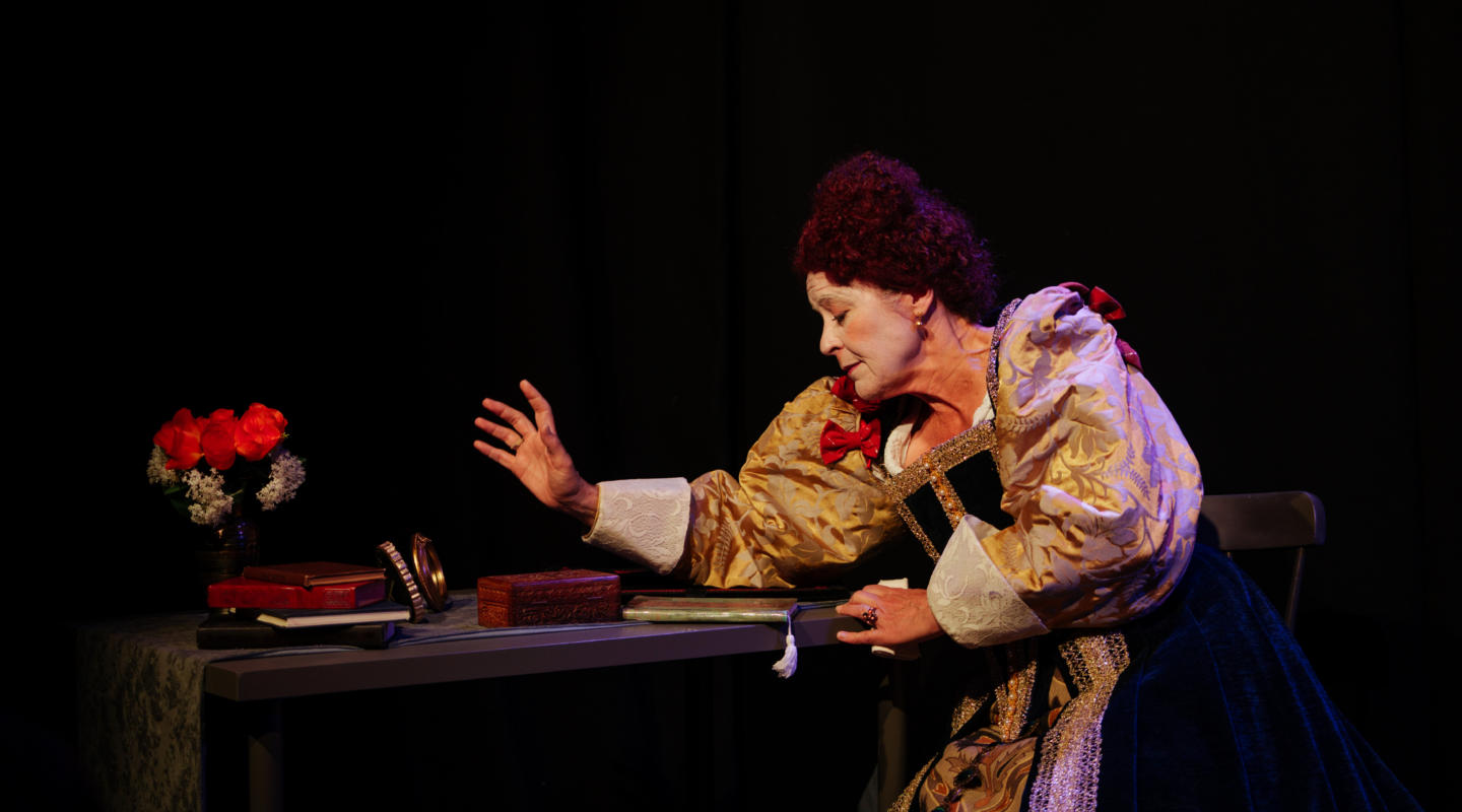 An actor in Elizabethan costume and white face makeup sits at a table with books, a mirror, and a vase of red and white flowers, reaching out with one hand.