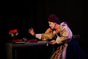 An actor in Elizabethan costume and white face makeup sits at a table with books, a mirror, and a vase of red and white flowers, reaching out with one hand.