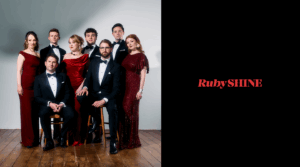 Eight people in formal attire pose for a group photo in a studio with a wooden floor; the right side displays the text "RubySHINE" on a black background.