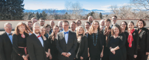 A group of adults dressed in formal attire stands outdoors in front of trees and distant mountains, smiling at the camera under a clear sky.