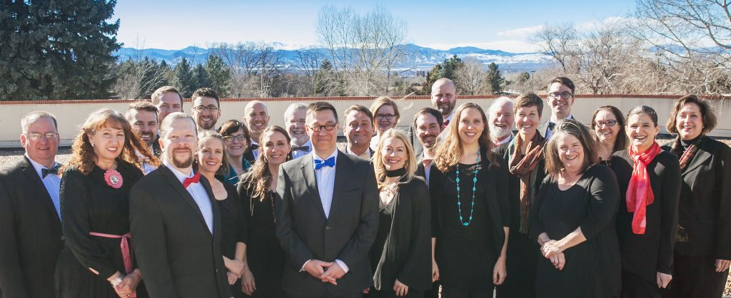 A group of adults dressed in formal attire stands outdoors in front of trees and distant mountains, smiling at the camera under a clear sky.