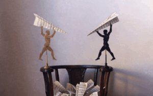 Two mannequin figures, one light and one dark, stand on a chair holding large paper airplanes made of sheet music, with more sheet music scattered below.