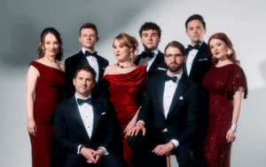 Eight members of the Colorado Choir in formal evening wear pose against a light background; two men are seated, while five others stand behind them on a wooden floor.