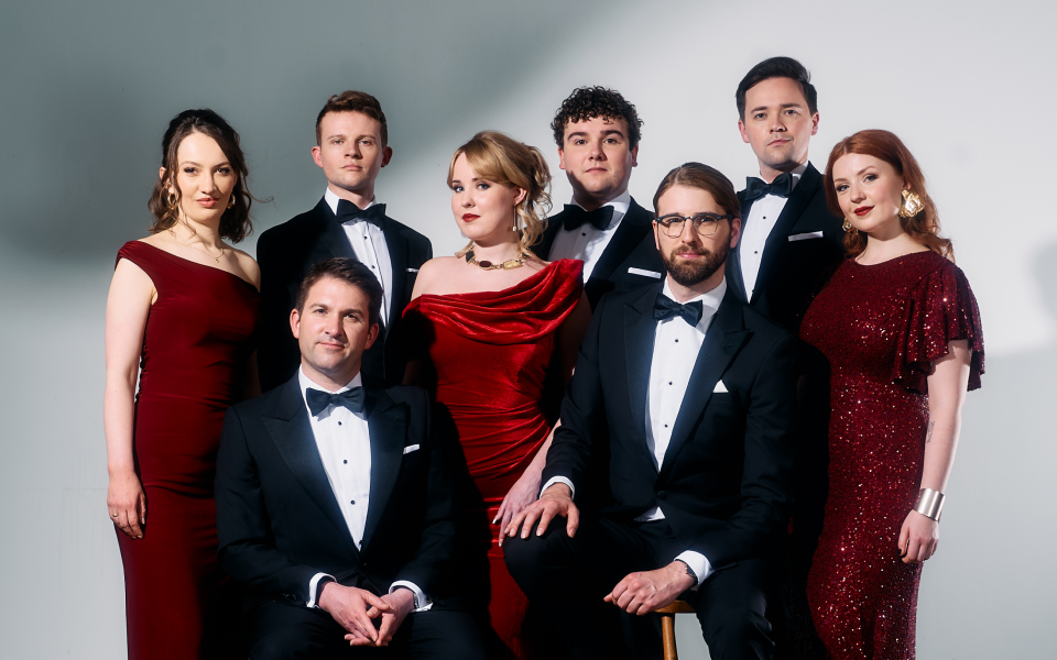 A group of four men in tuxedos and four women in red formal dresses pose together against a plain background.