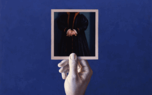 A hand holds a photo featuring a person in a dark coat with their head cropped out, against a solid blue background.