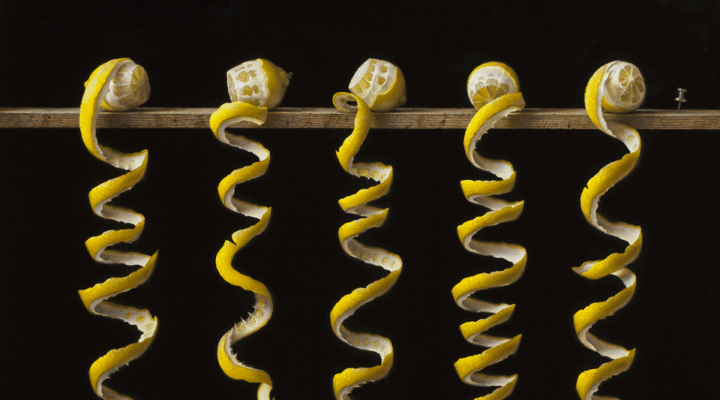 Five lemons with long, continuous peels spiral downward from a wooden rod against a black background.