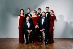 Eight adults in formal evening attire, with five standing and three seated on chairs, pose against a plain backdrop on a wooden floor.
