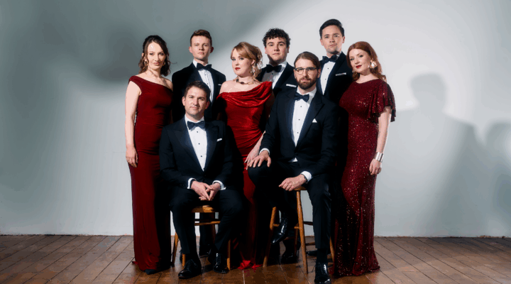 Eight adults in formal evening attire, with five standing and three seated on chairs, pose against a plain backdrop on a wooden floor.