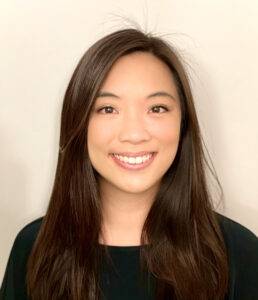A woman with long, straight brown hair and a black top smiles at the camera against a plain light background.