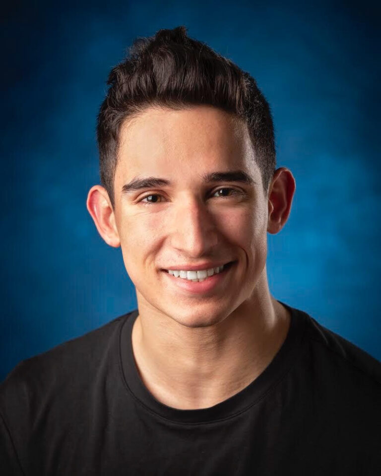 Tenor Nikhil Harle, a young man with short dark hair and a black t-shirt, smiles at the camera against a blue background.