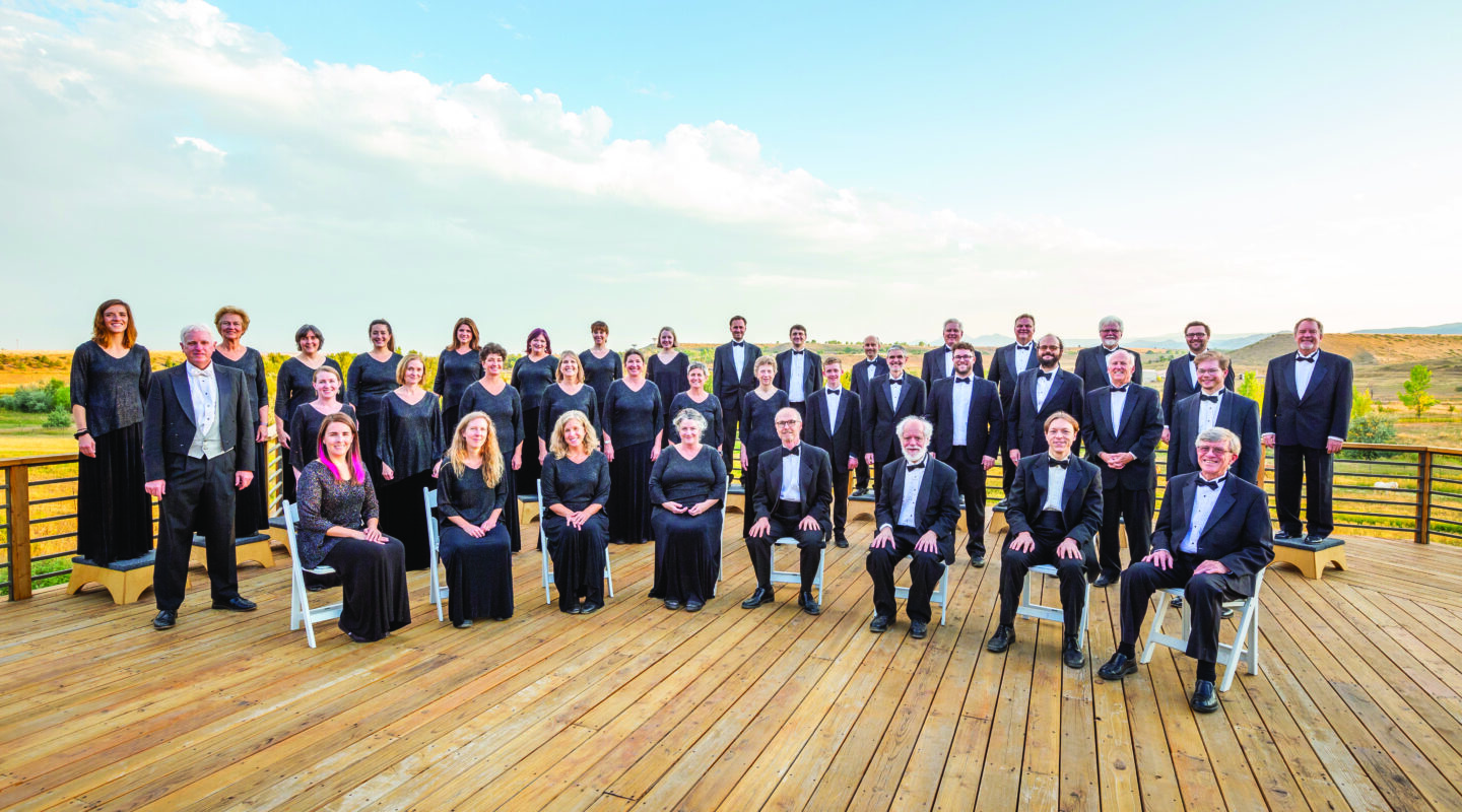 A large group of men and women dressed in formal black attire pose on a wooden deck outdoors, celebrating 40 Years of the Ars Nova Singers with a scenic sky and landscape in the background—truly music beyond words.