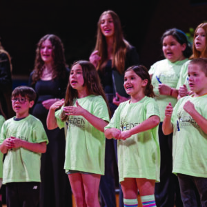 A group of children in light green shirts stands singing or performing on stage, celebrating 40 Years of Music Beyond Words, with a few adults in dark clothing—possibly Ars Nova Singers—behind them.